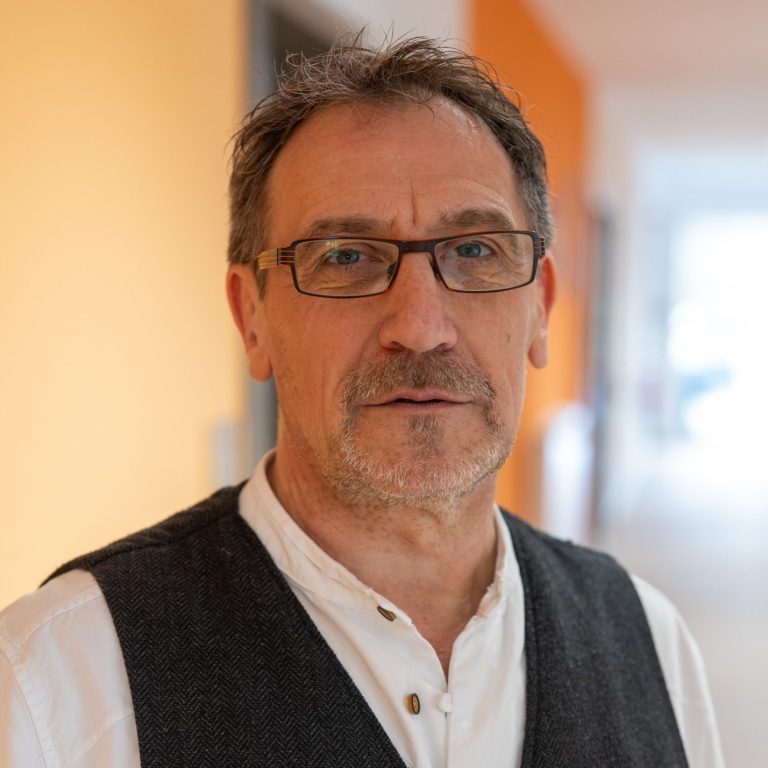 Mann mit Brille und Weste, steht in einem hellen Flur mit orangefarbener Wand.