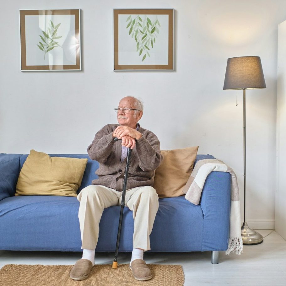 Ein älterer Mann sitzt auf einem blauen Sofa mit einem Stock, umgeben von Pflanzenbildern.