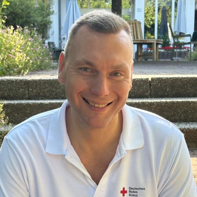 Mann in blauer Polo-Uniform vor einer grünen, blühenden Landschaft.