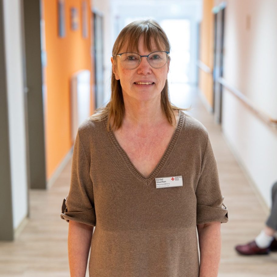 Frau mit Brille und braunem Oberteil lächelt in einem Flur mit orangefarbenen Wänden.