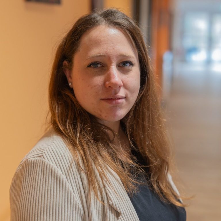 Porträt einer Frau mit langen, braunen Haaren in einem Flur mit orangefarbener Wand.