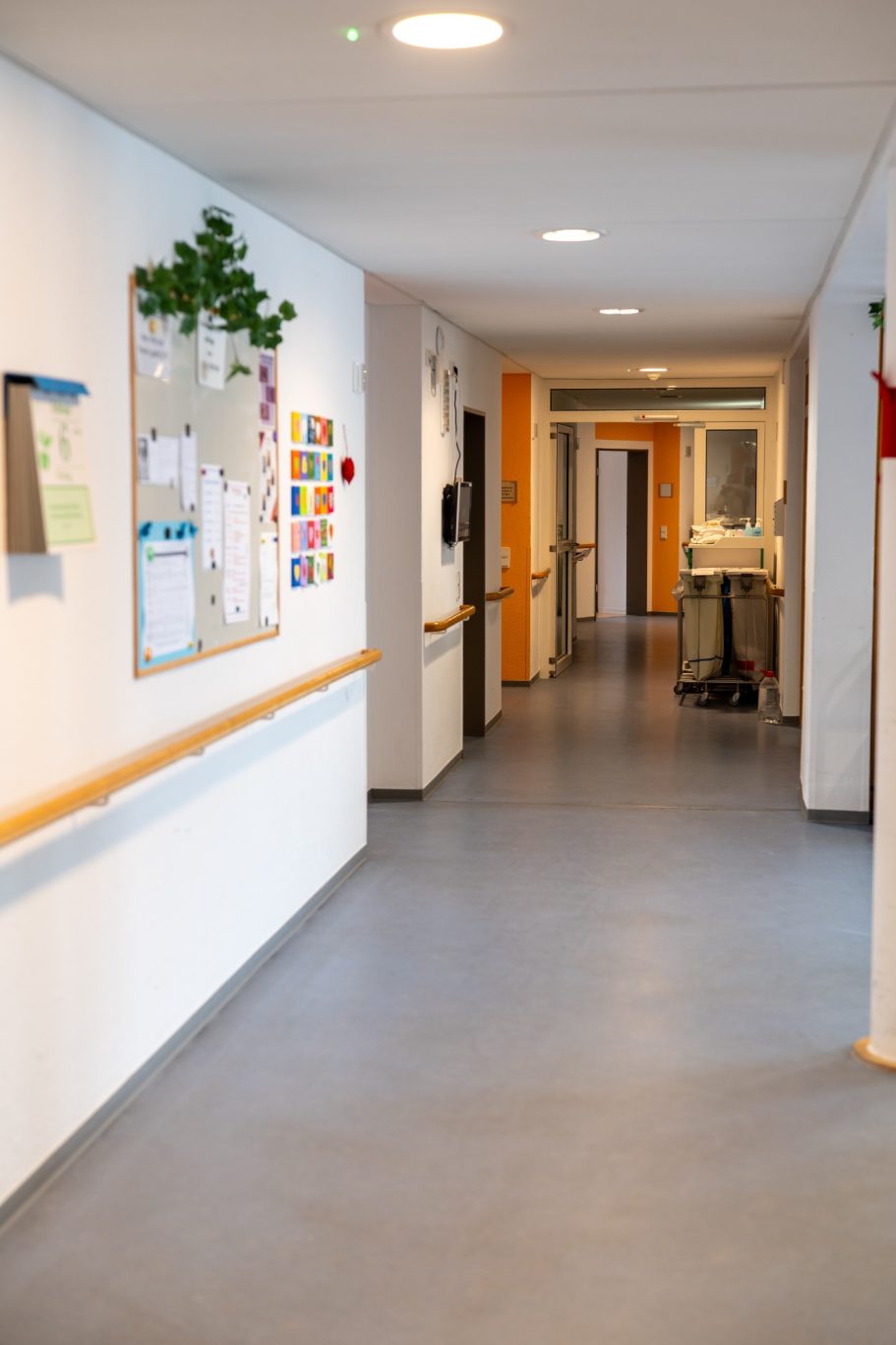 Heller Krankenhausflur mit Wänden voller Informationen und einem Überblick auf Patientenzimmer.