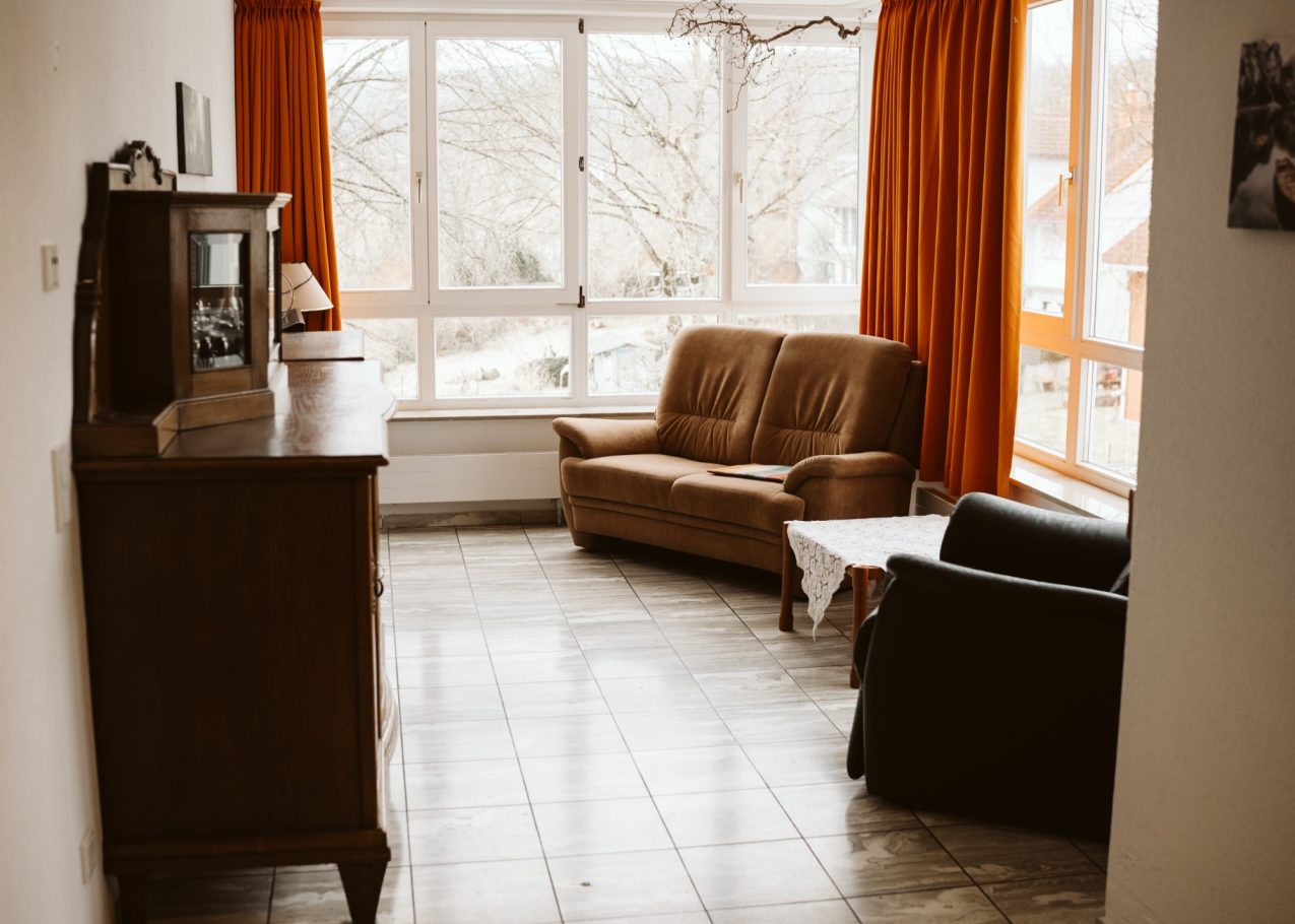 Gemütliches Wohnzimmer mit zwei Sofas, einem Tisch und großen Fenstern mit oranges Vorhänge.