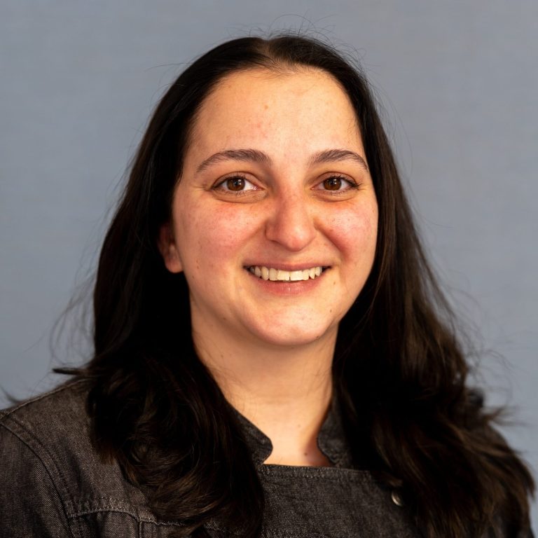 Lächelnde Person mit langen, dunklen Haaren und blauer Jacke vor grauem Hintergrund.
